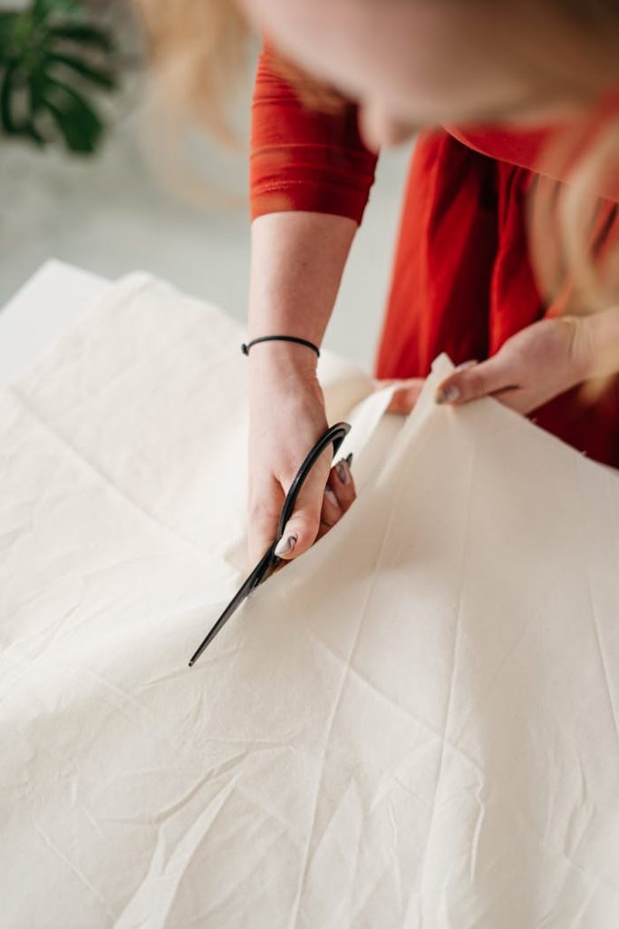 person-in-red-long-sleeve-shirt-holding-black-and-silver-scissors-3738075 Focused seamstress cutting white cotton fabric with scissors, showcasing zero waste fashion.